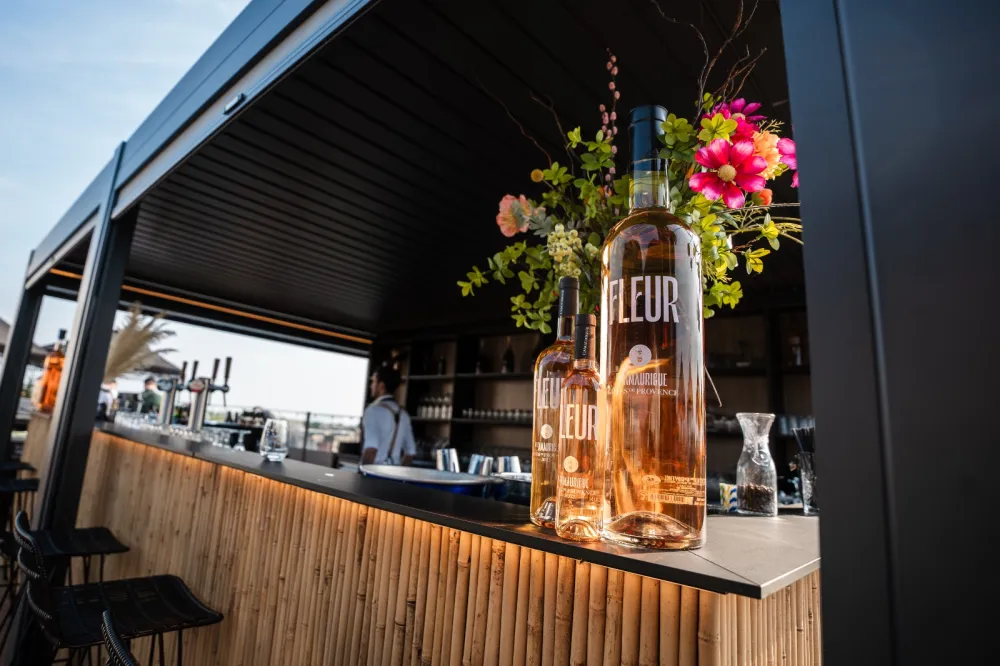 Flessen roséwijn staan op een bar met een buitenkant van bamboe. Op de achtergrond is een barman te zien die drankjes klaarmaakt. Felgekleurde bloemen sieren de bar en geven het tafereel een vleugje kleur. Er zijn zitplaatsen buiten in de buurt.