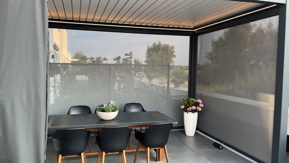 Een modern buitenterras heeft een zwarte tafel omringd door zwarte stoelen onder een pergola. Een witte plantenbak gevuld met bloemen staat op de tafel en een grote witte vaas met roze bloemen staat bij de hoek. De muren zijn gemaakt van doorzichtig gaas.