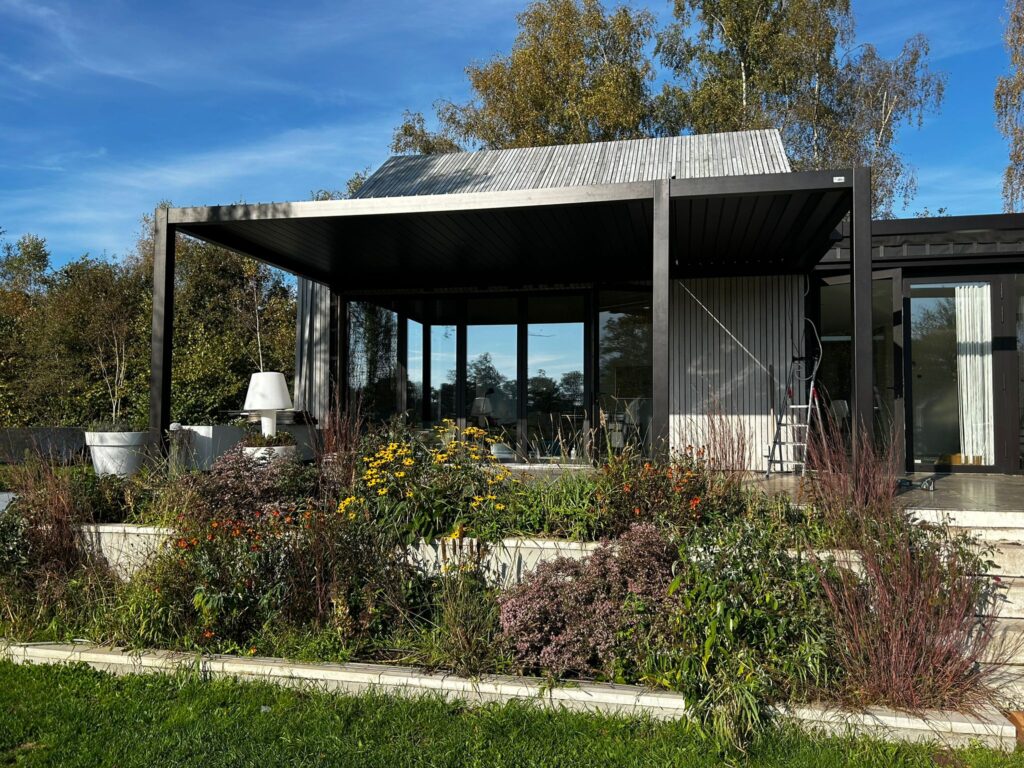 Een modern huis met grote glazen deuren is omgeven door een weelderige tuin vol met verschillende bloemen en planten. Het gebouw heeft een metalen dak en een patio met zitplaatsen. Bomen zijn zichtbaar op de achtergrond onder een helderblauwe lucht.