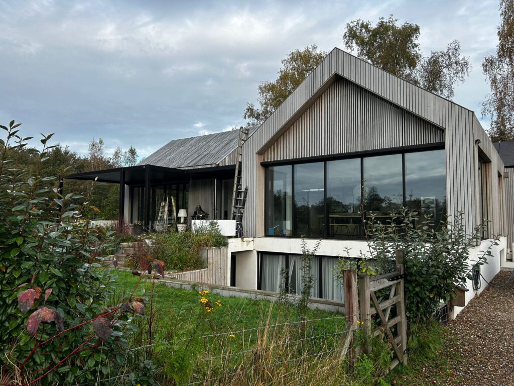 Modern huis met een gegolfde metalen buitenkant en grote ramen omgeven door groen. Het gebouw heeft twee verbonden delen met schuine daken. Een tuin met verschillende planten en een houten hek zijn zichtbaar op de voorgrond.