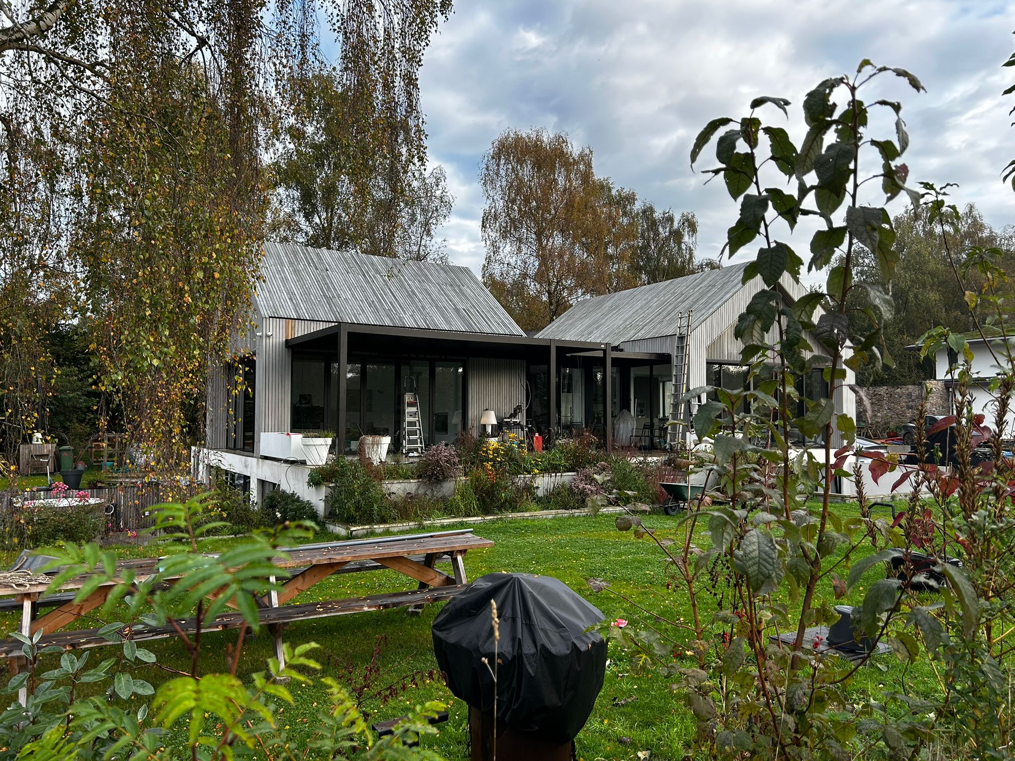 Een modern, tweedelig zadeldakhuis met metalen bekleding is omgeven door bomen en een grasveld. Op de voorgrond staat een picknicktafel en een tuin met verschillende planten. De lucht is bewolkt, wat het minimalistische ontwerp van het huis versterkt.