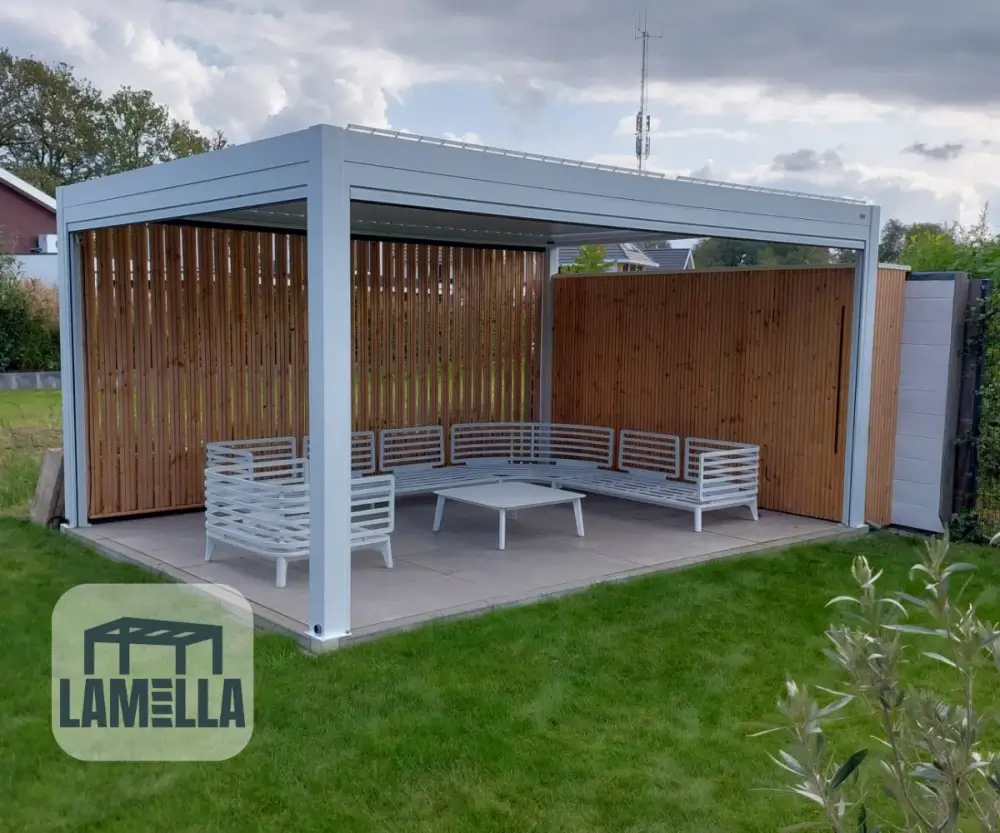 Een moderne, witte pergola met een plat dak en houten lattenwanden staat op een patio. Het heeft een witte L-vormige sectionele bank en een glazen salontafel. De pergola staat in een grasachtige achtertuin. Het Lamella-logo is zichtbaar in de linkerbenedenhoek.