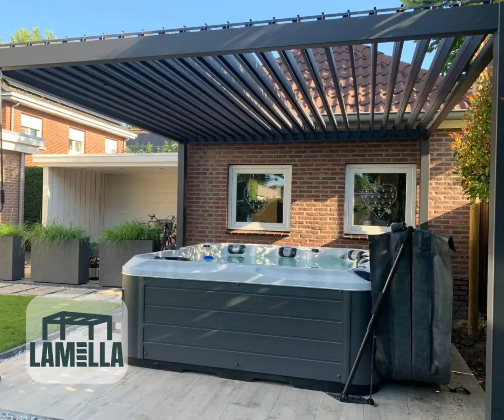 Een moderne patio buiten heeft een hottub onder een metalen pergola met verstelbare lamellen. De omgeving bestaat uit een bakstenen muur met twee ramen en potplanten. Het logo Lamella is zichtbaar in de linkerbenedenhoek.