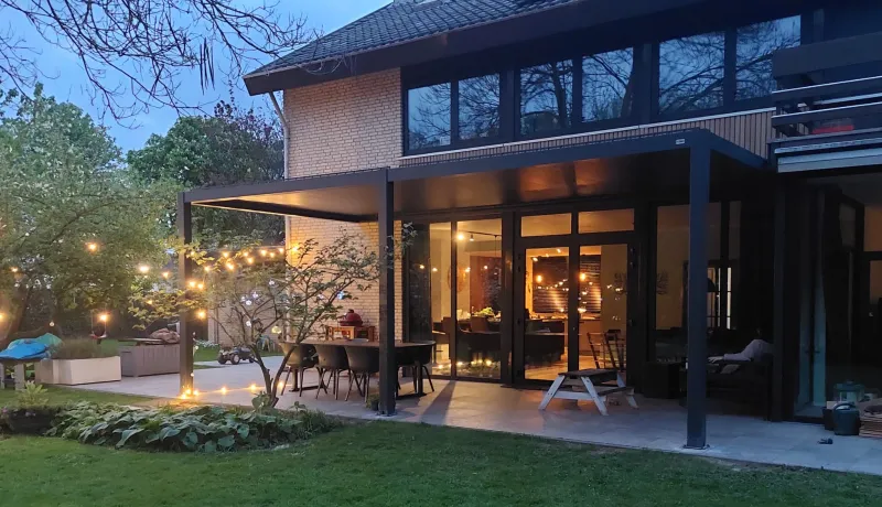 Een modern huis met een groot terras en lichtsnoeren. Het overdekte buitengedeelte is ingericht met een eettafel en stoelen. Weelderig groen omringt het huis en binnenverlichting creëert een warme ambiance.