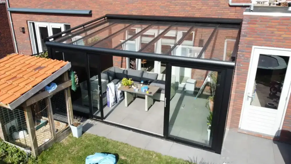 Een moderne glazen serre die aan een bakstenen huis vastzit, met zwarte frames. Binnen is er een zithoek met een bankstel en een tafel. De omgeving bestaat uit een klein houten schuurtje en een groen gazon met tuinspullen.