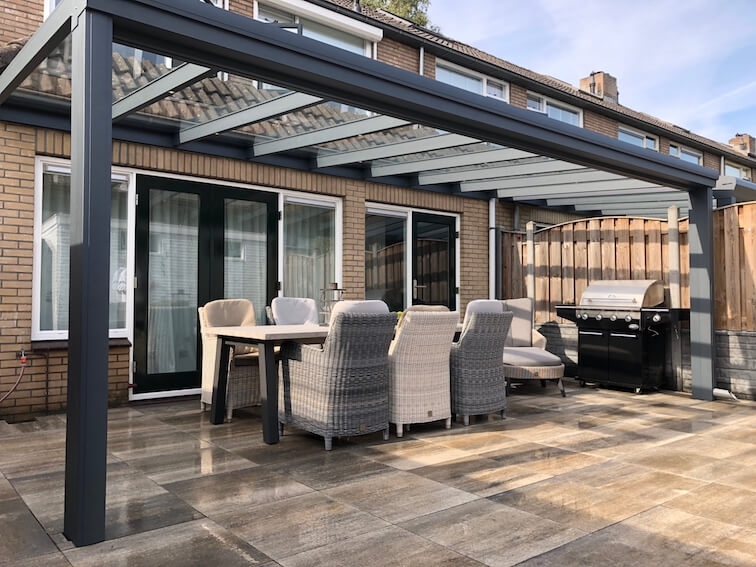 Een patio met een Terrasoverkapping glas bedekt een eethoek met een rechthoekige tafel en zes stoelen met kussens. In de buurt staat een zwarte barbecue. Het gebied is geplaveid met grote tegels en op de achtergrond staat een bakstenen huis met glazen schuifdeuren.