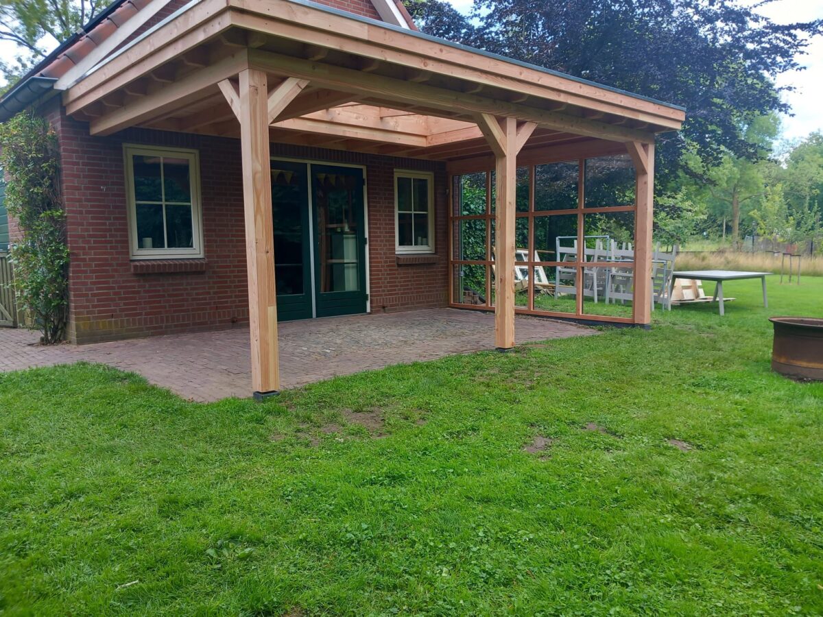 Ein Haus mit Backsteinfassade verfügt über die Houten overkapping Classic, eine hölzerne Terrassenüberdachung mit Gitterseiten. Eine Glastür führt auf die Terrasse mit Tisch und Stühlen. Der Bereich umfasst einen grünen Rasen und Bäume, in der Nähe befindet sich eine große zylindrische Feuerstelle.