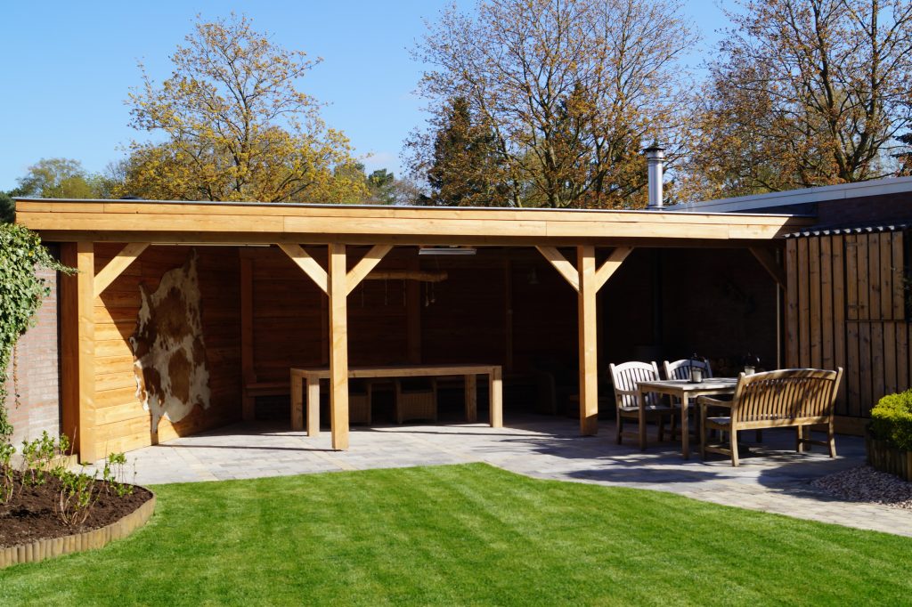 Een Houten overkapping Classic staat in een achtertuin met groen gras, met een lange tafel en banken eronder. Extra houten tuinmeubilair staat op het stenen terras ernaast. Bomen op de achtergrond vertonen herfstbladeren.