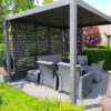 Een moderne zithoek in de buitenlucht met een grijze pergola met lamellenpanelen. Binnen staan rieten stoelen met kussens en een salontafel. De patio is omgeven door weelderig groen en een boom met rode bladeren, met een bakstenen gebouw op de achtergrond.