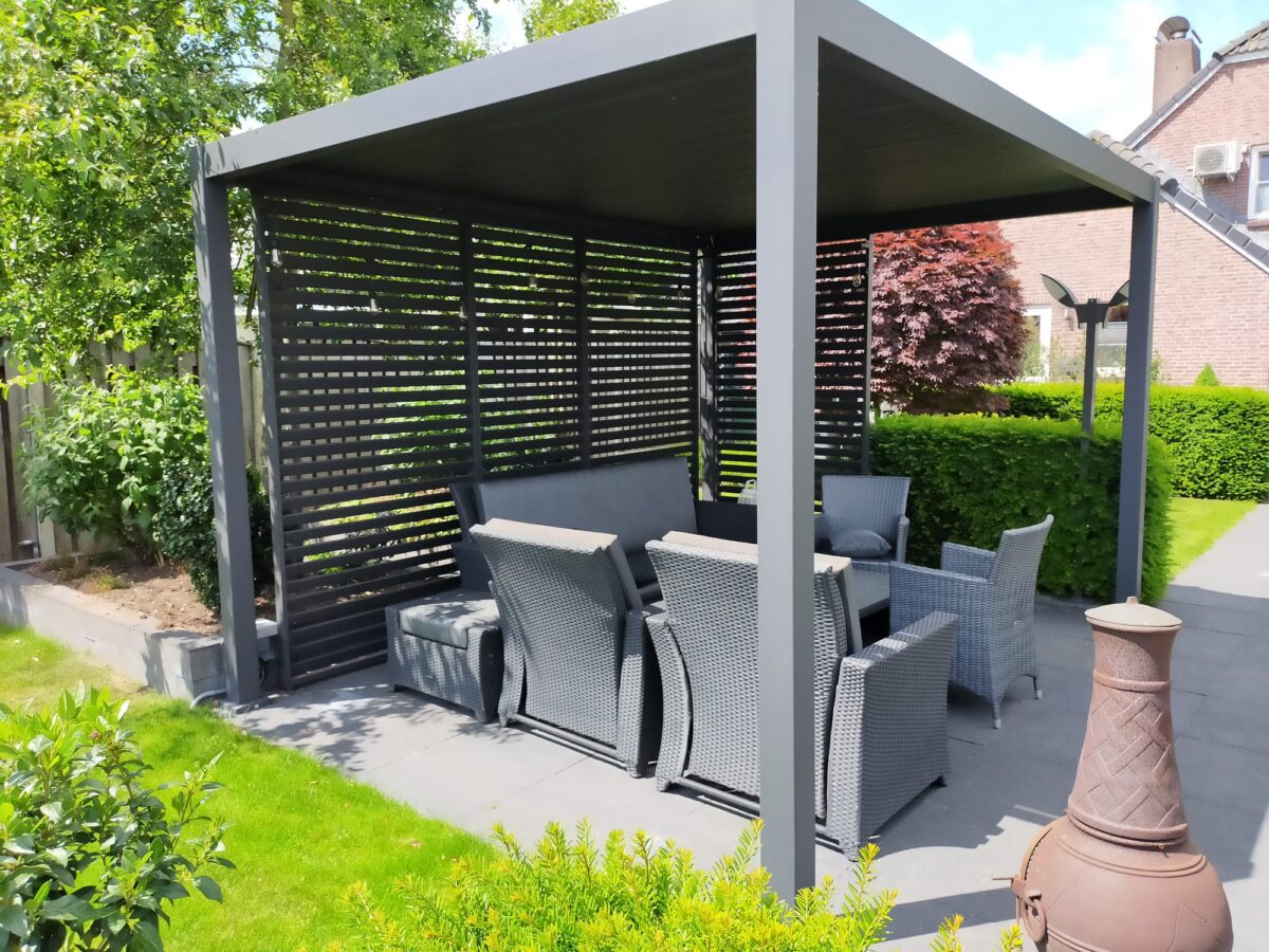 Een moderne zithoek in de buitenlucht met een grijze pergola met lamellenpanelen. Binnen staan rieten stoelen met kussens en een salontafel. De patio is omgeven door weelderig groen en een boom met rode bladeren, met een bakstenen gebouw op de achtergrond.