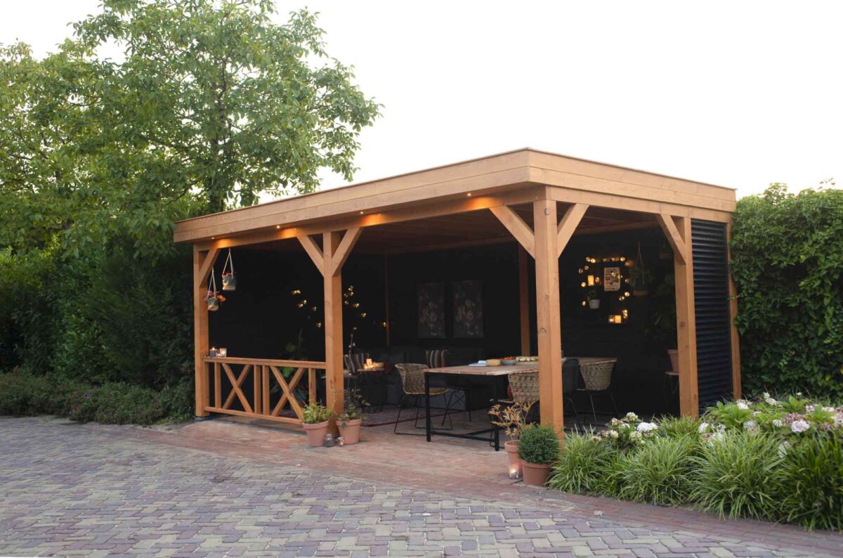 Een Houten overkapping Classic staat in de tuin met een plat dak, verlicht door lichtsnoeren die de tafel en stoelen binnen verlichten. Potplanten en weelderig groen omringen het, terwijl een bakstenen patio charme toevoegt aan de voorgrond.