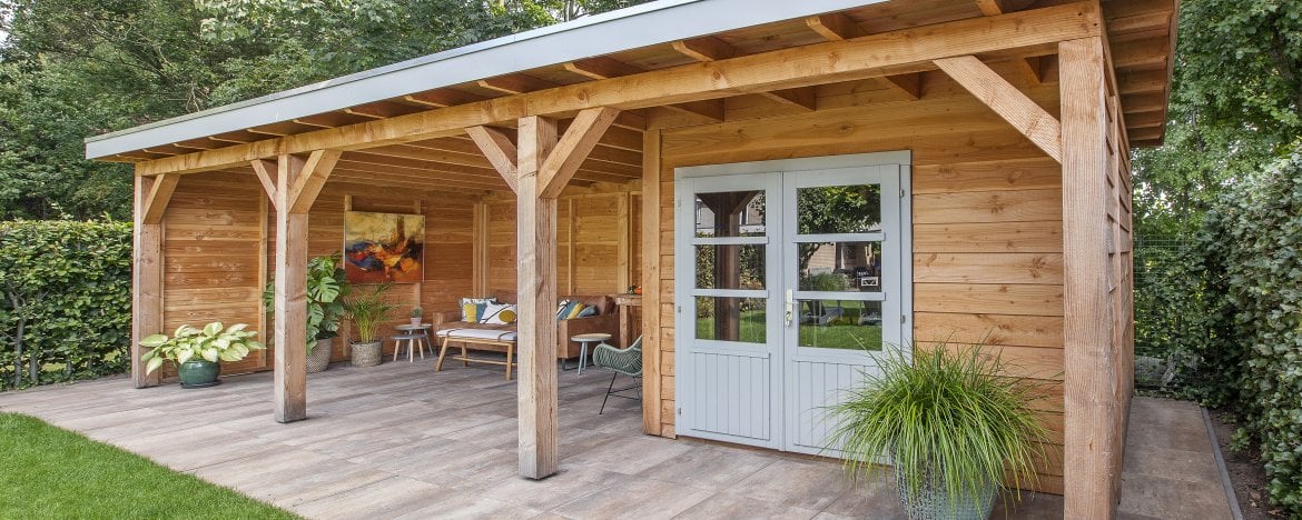 Een houten tuinhuisje met een schuin dak en dubbele deuren bevindt zich onder de charmante Houten overkapping Deluxe, met een kleine veranda met een bank en kussens. Potplanten sieren de veranda en de entree, omgeven door weelderig groen.