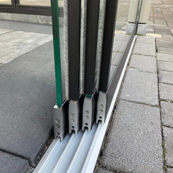 Close-up van drie glazen schuifpanelen met metalen frames op een rails, geplaatst op een betegeld buitenoppervlak.