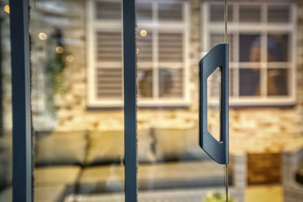 Close-up van een glazen deurgreep met een wazige achtergrond die een gezellig terras laat zien. Het terras heeft rieten meubels en een bakstenen muur met luiken voor de ramen, wat een warme en uitnodigende sfeer creëert.