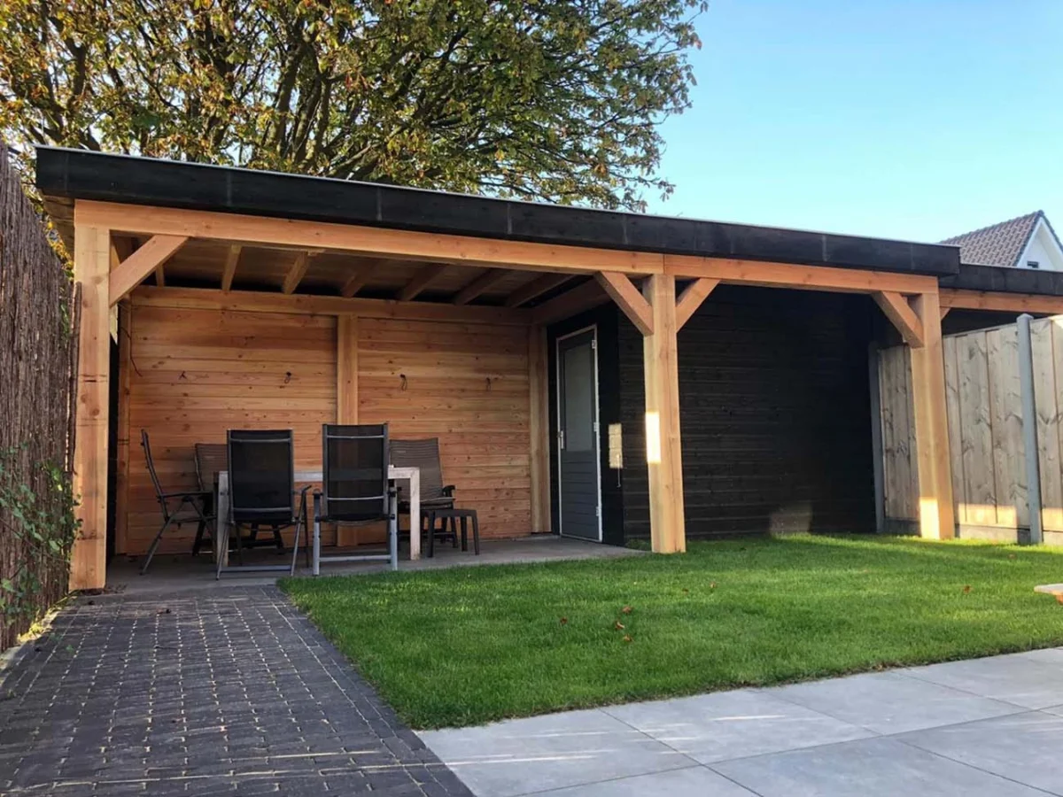 De Deluxe Houten overkapping is een houten tuinhuis met zwarte wanden en een licht houten frame. Het bevat een tafel met vier stoelen, is omgeven door tuingras en een geplaveid pad, en heeft een grote boom op de achtergrond.