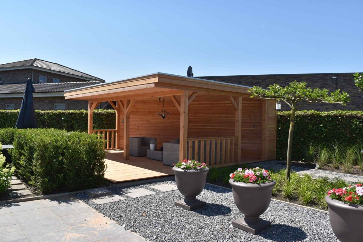 Een Houten overkapping Classic houten prieel met zitgelegenheid siert de achtertuin, omlijst door nette heggen en een stenen terras. Het beschikt over drie grote plantenbakken gevuld met kleurrijke bloemen, allemaal onder een helderblauwe lucht.