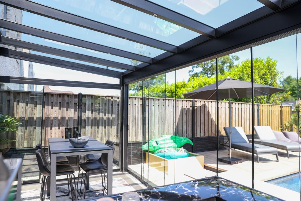 Licht en zonnig modern terras met Terrasoverkapping glas, met glazen wanden en dak, een eettafel met stoelen, ligstoelen, groene zitzakken, omgeven door een houten hek en groen buiten voor een ontspannen sfeer.