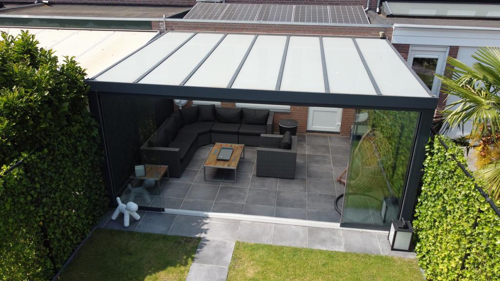 Een modern buitenterras met het Terrasoverkapping glazen dak en donker metalen frame, met een hoekbank en tafel op betegelde vloer. Omgeven door heggen, een palmboom en een grasveld met een witte teddybeersculptuur.