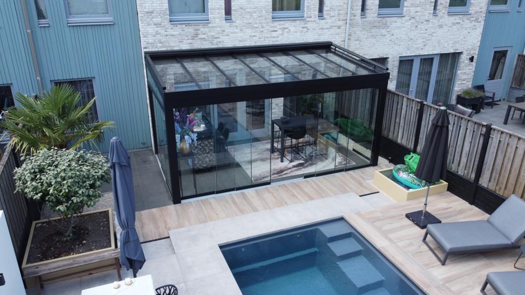 Een moderne achtertuin heeft een klein zwembad en een houten terras, met een Terrasoverkapping glas dat de patio omsluit met een eettafel en potplanten. Buitenstoelen en parasols zijn rond de ruimte gerangschikt, allemaal binnen een houten hek.