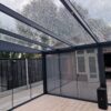 Moderne patio voorzien van Terrasoverkapping glas, met transparant dak en glazen wanden met een gedeeltelijk open deur naar een omheind buitengebied. Binnen een betegelde vloer en potpalm. Bewolkte lucht gezien door het natte glazen dak.