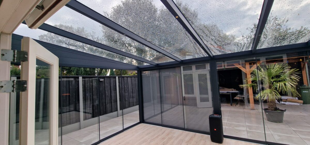 Moderne patio voorzien van Terrasoverkapping glas, met transparant dak en glazen wanden met een gedeeltelijk open deur naar een omheind buitengebied. Binnen een betegelde vloer en potpalm. Bewolkte lucht gezien door het natte glazen dak.