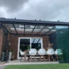 Een moderne eethoek in de buitenlucht met de Terrasoverkapping kubus heeft een glazen dak en muren die aan een bakstenen huis zijn bevestigd. Het bevat een lange houten tafel, witte stoelen, decoratieve planten en is omgeven door groen onder een bewolkte lucht.