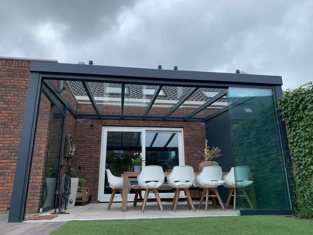 Een moderne eethoek in de buitenlucht met de Terrasoverkapping kubus heeft een glazen dak en muren die aan een bakstenen huis zijn bevestigd. Het bevat een lange houten tafel, witte stoelen, decoratieve planten en is omgeven door groen onder een bewolkte lucht.