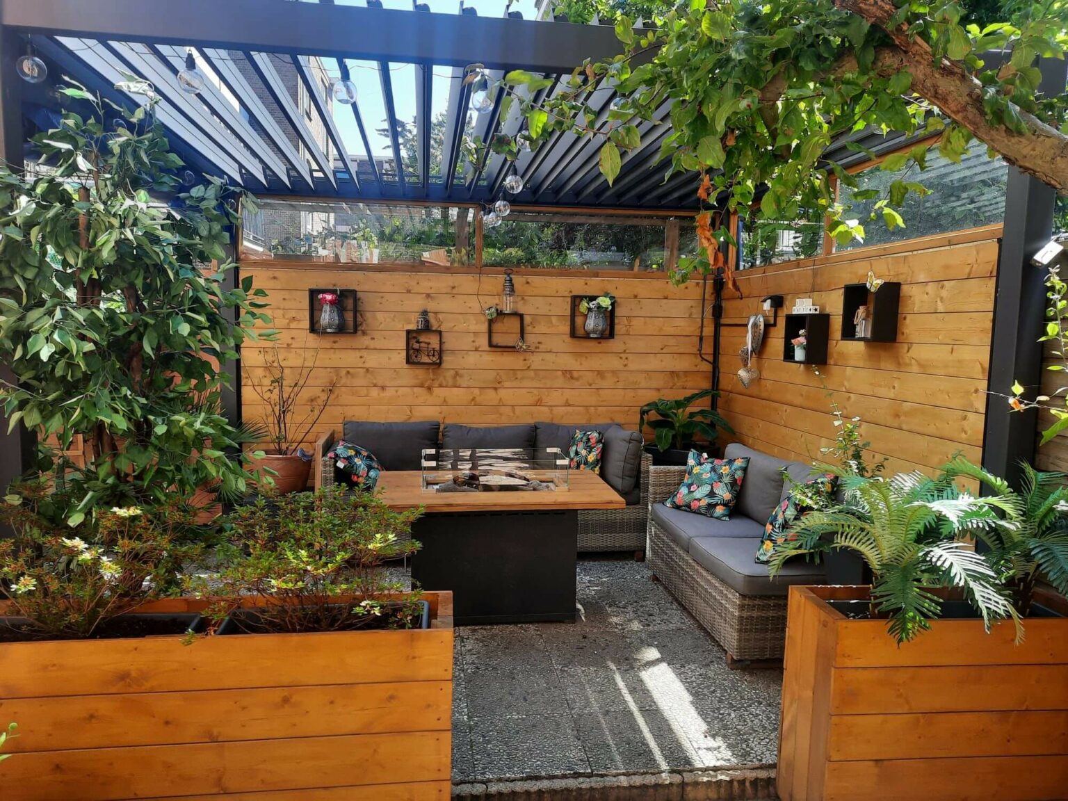 Gezellige patio in een kleine tuin met een pergola met latten, houten wanden, een L-vormige bank met kussens en een salontafel in het midden. De inrichting bestaat uit levendige kussens en hangplanten. Omringd door weelderig gebladerte in houten plantenbakken. Heldere, uitnodigende sfeer. lamellen overkapping.