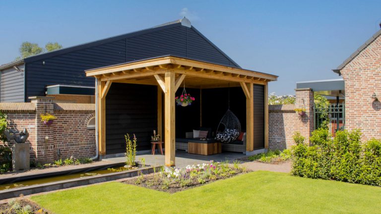 Die Houten overkapping Classic steht in einem Hinterhof mit einer schwarz gestrichenen Wand und Ziegelelementen, ergänzt durch Gartenmöbel und einen Hängesessel. Im Vordergrund gedeiht ein Garten mit Teich, Pflanzen und Blumen unter dem klaren blauen Himmel.