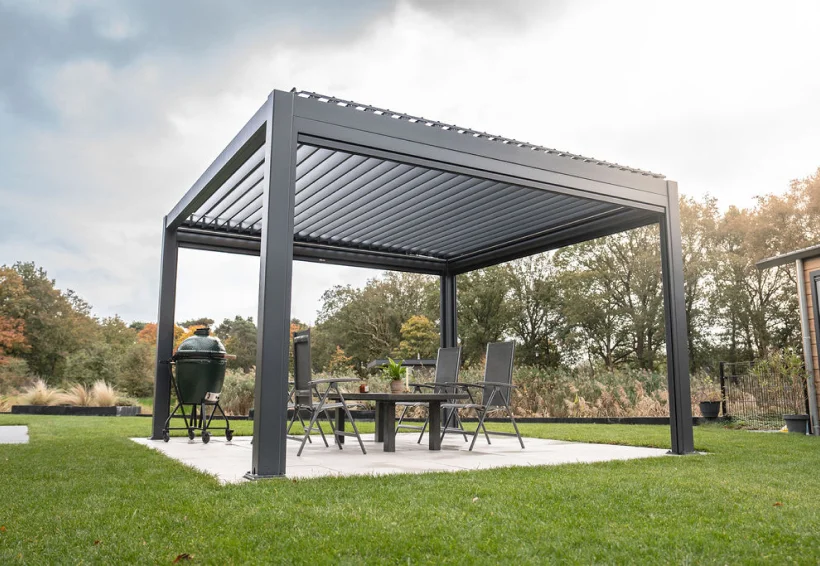 Een moderne buitenprieel met een strak metalen frame staat op een terras. Daaronder staat een eettafel met stoelen en decoratieve planten. Rondom het prieel ligt een goed onderhouden gazon en bomen op de achtergrond onder een bewolkte lucht.