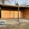 Houten schuur met een plat dak en open voorkant, ondersteund door donkere metalen balken. De grond is zanderig en er zijn kale bomen en een huis zichtbaar op de achtergrond. Een LAMELLA-logo staat in de linkerbenedenhoek.