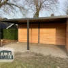 Een moderne houten schuilplaats met een plat dak en drie muren staat in een tuin. De constructie heeft een open voorkant en wordt ondersteund door vier metalen palen. Een logo met de tekst Lamella wordt in de linkerbenedenhoek weergegeven. Bomen en een heg staan op de achtergrond.