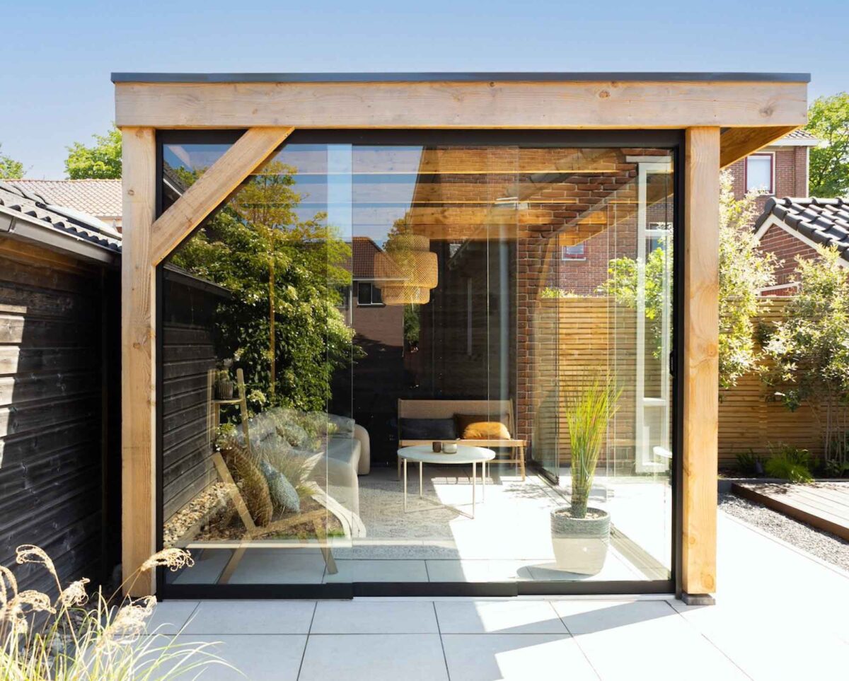 Een moderne tuinkamer met grote glaspanelen en een houten balkenstructuur die lijkt op de Houten overkapping Classic, heeft een gezellig interieur met een witte bank, ronde tafel en kamerplanten. Het zonlicht stroomt naar binnen en verlicht het omringende groen en het nabijgelegen huis.