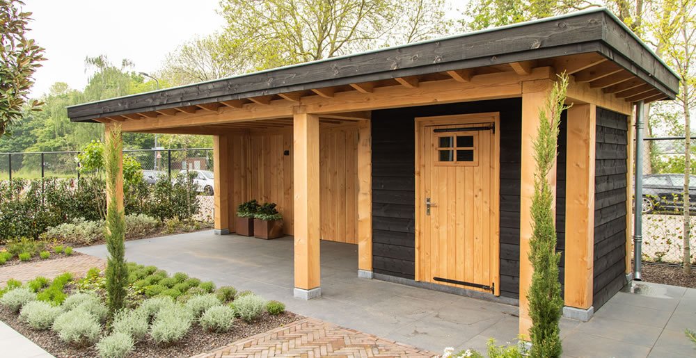 Een houten schuur met een kleine overhang en een enkele deur, met zwarte en natuurlijke houten panelen. Het is omgeven door een netjes aangelegde tuin met kleine struiken en bomen. De lucht is helder en er is een metalen hek zichtbaar op de achtergrond.