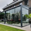 Modern huis met een glazen Terrasoverkapping glas, met een hangende eierstoel, rood-witte meubels en palmbomen in potten op de tegels. De achtergrond heeft een netjes bijgehouden gazon en een grote bakstenen gevel met ramen.