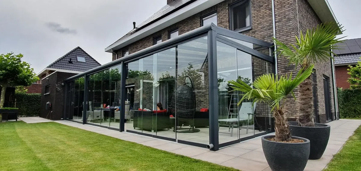 Modern huis met een glazen Terrasoverkapping glas, met een hangende eierstoel, rood-witte meubels en palmbomen in potten op de tegels. De achtergrond heeft een netjes bijgehouden gazon en een grote bakstenen gevel met ramen.