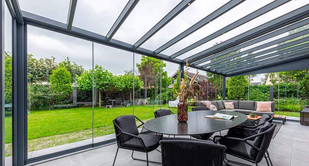 De Terrasoverkapping glas omsluit een moderne patio met een ronde zwarte tafel en zes stoelen, versierd met een gedroogd bloemenarrangement. Een grijze bank met kussens staat in de hoek, met uitzicht op een weelderige groene tuin vol met verschillende bomen.