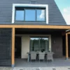 Modern huis met een schuin dak en een groot raam op de bovenverdieping. Een overdekte patio heeft een rieten eethoek onder een houten pergola. De buitenkant heeft contrasterende donkere bakstenen en beige muren. Een hek en wat groen zijn zichtbaar aan de zijkant.