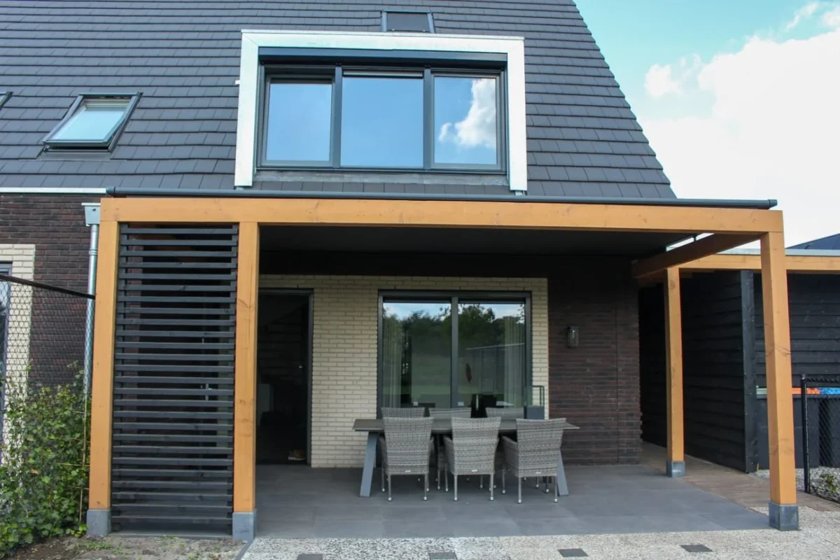 Modern huis met een schuin dak en een groot raam op de bovenverdieping. Een overdekte patio heeft een rieten eethoek onder een houten pergola. De buitenkant heeft contrasterende donkere bakstenen en beige muren. Een hek en wat groen zijn zichtbaar aan de zijkant.