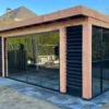 Een moderne tuinkamer met grote glazen panelen en houten accenten. De structuur heeft zwarte lattenbekleding, omgeven door een geplaveid terras. Een houten picknicktafel is zichtbaar op de achtergrond.
