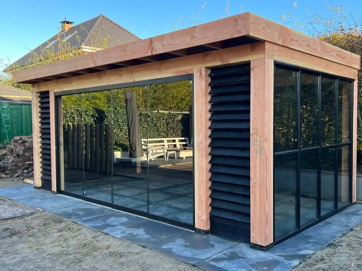 Een moderne tuinkamer met grote glazen panelen en houten accenten. De structuur heeft zwarte lattenbekleding, omgeven door een geplaveid terras. Een houten picknicktafel is zichtbaar op de achtergrond.