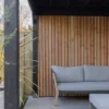 Een moderne zithoek buiten heeft een grijze, gestoffeerde bank tegen een verticale houten lattenwand. Potplanten flankeren de ruimte en een betegelde vloer loopt door onder een donkere pergola, wat een stijlvolle en gezellige sfeer creëert.