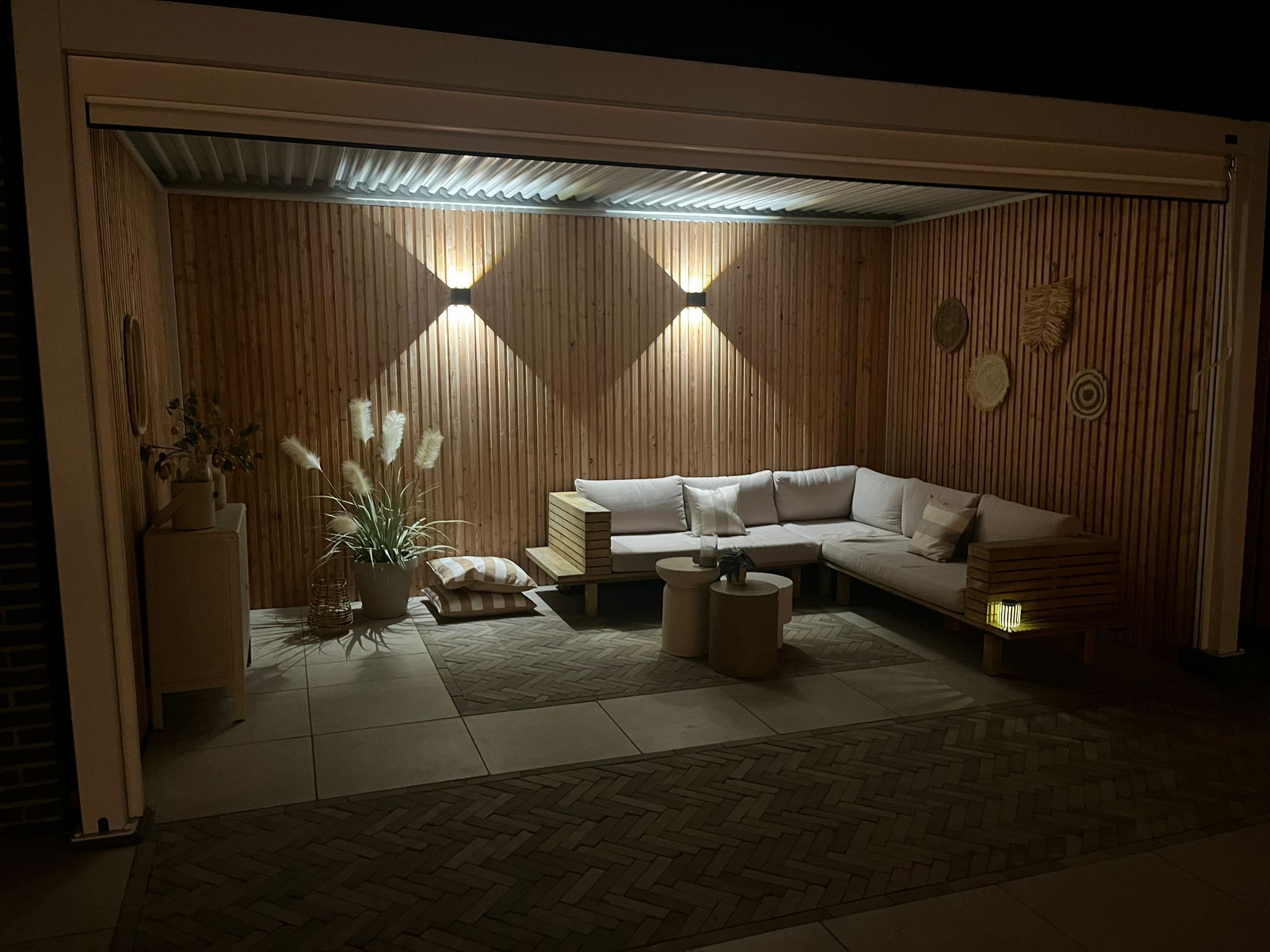 Een gezellige loungeplek in de buitenlucht s avonds heeft een houten muur met twee wandlampen, een hoekbank met beige kussens en een kleine ronde tafel. Geaccentueerd door een potplant en geweven wandkunst, is de ruimte uitnodigend omsloten met een lamellen overkapping.