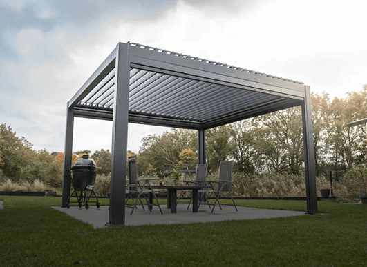 Op een betonnen terras in een grasveld staat een moderne metalen pergola met een lamellendak. Er staan een eettafel, stoelen en een barbecue, omringd door bomen en groen onder een gedeeltelijk bewolkte hemel.