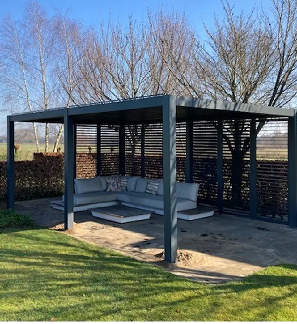 Een moderne buitenpergola met een dak met lamellen en zijkanten, met een grijze slaapbank met kussens, staat op een stenen patio, omringd door gras en bladerloze bomen onder een strakblauwe hemel.