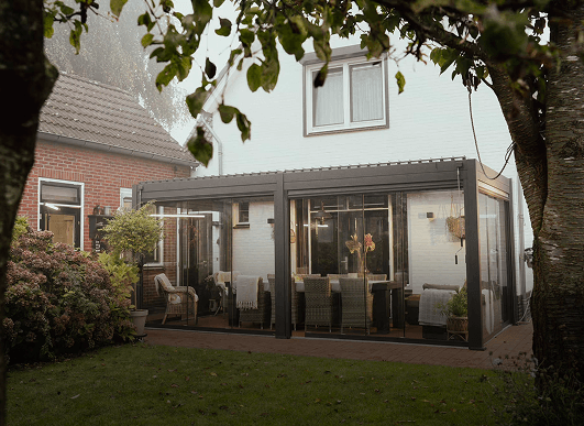 Een overdekte patio grenzend aan een wit huis, met glazen wanden, rieten stoelen, een eettafel en potplanten. Het gebied is omgeven door groen en bomen in een achtertuin.