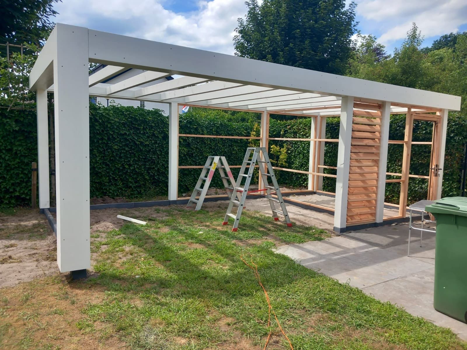 Een gedeeltelijk gebouwde lamellenoverkapping of tuinhuisje met een wit frame en houten latten; binnenin staan twee ladders en op het gras onder een gedeeltelijk bewolkte hemel staat een groene bak met verlengsnoer.