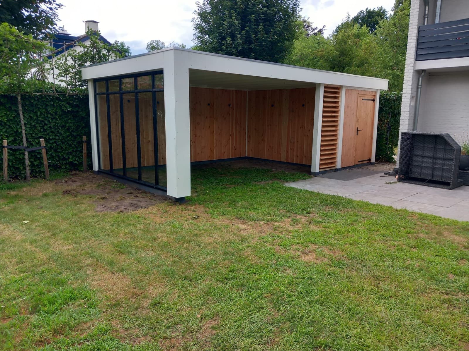 Een modern houten tuinhuisje met een wit dak, glaspanelen aan één kant, een klein deurtje en een overdekte open ruimte met een stijlvolle lamellenoverkapping, gelegen op een gazon naast een terras en omgeven door groen.