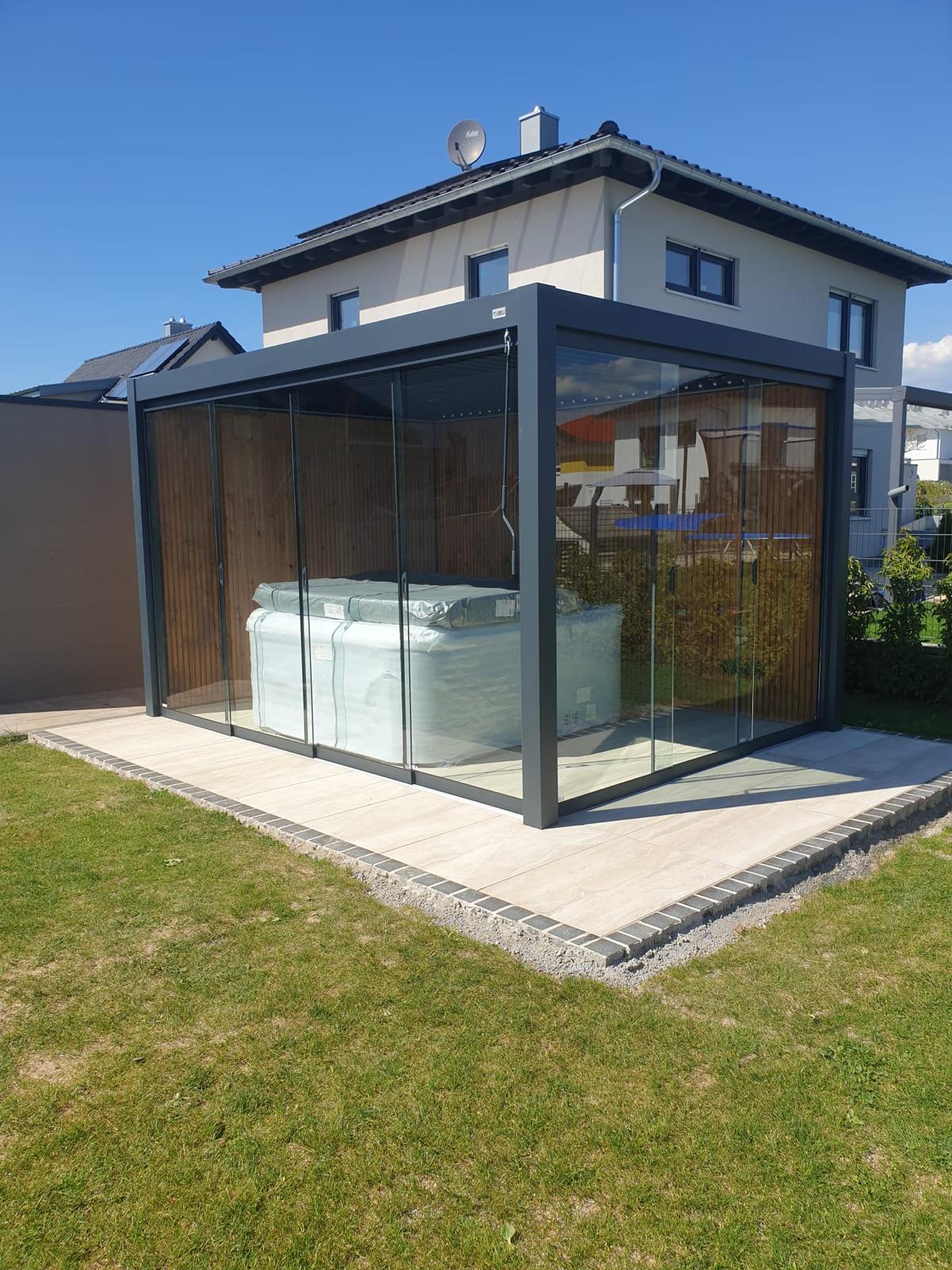 Een moderne overkapping van glas en metalen lamellen staat op een betegeld terras in een met gras begroeide achtertuin en biedt beschutting aan iets dat overdekt is. Op de achtergrond staat een huis met twee verdiepingen onder een helderblauwe lucht.