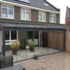 Een moderne woning met een terrasoverkapping op maat met een glazen omheining, potplanten, tegelvloer en houten schutting. De buitenkant heeft grote ramen en een donker dak.